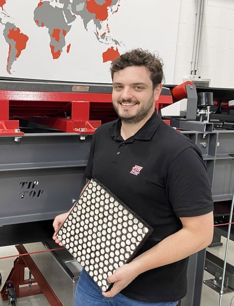 A smiling man holding a perforated metal sheet lined with REMALOX rubber material, standing in front of a large press machine in an industrial workspace.